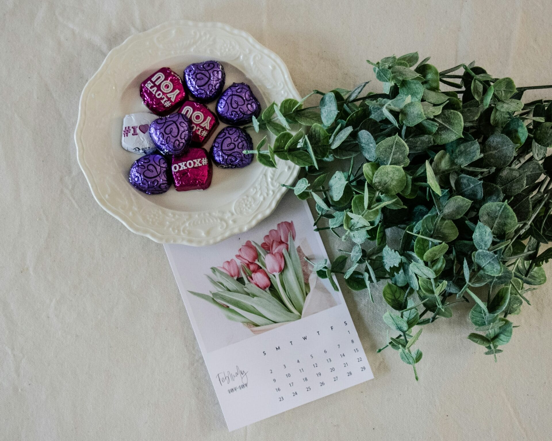 Green eucalyptus stems next to a ceramic bowl with chocolate hearts, each wrapped in purple or red foil